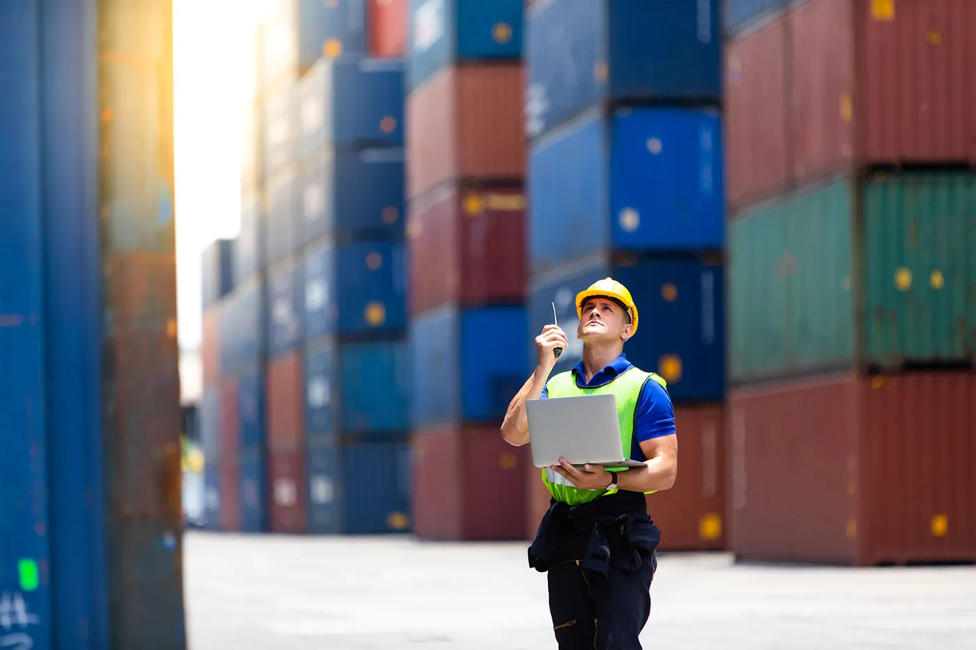 Worker wearing high vis and a hard hat holding a laptop and looking up at shipping containers